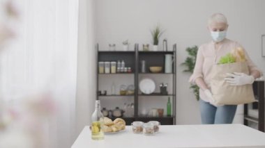 Yüzü maskeli ve eldivenli kadın marketten eve geliyor, kilitlenmekten memnun değil.