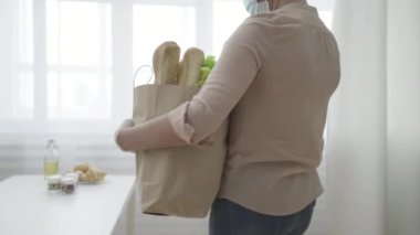 Koruyucu maskeli ve eldivenli bir kadın eve market poşeti getiriyor, kilitlenmekten yorulmuş.