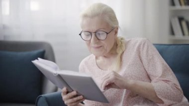 Yaşlı kadın kitap okurken torun akıllı telefonla oynuyor, aile dinleniyor.