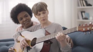 Siyahi bir kadın, bayan arkadaşına gitar öğretiyor. Çiftler birlikte hobi yapıyor.