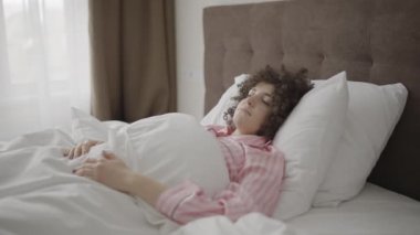 Genç bir kadın rahat bir yatakta uyanıyor, ellerini uzatıyor, hayattan zevk alıyor.