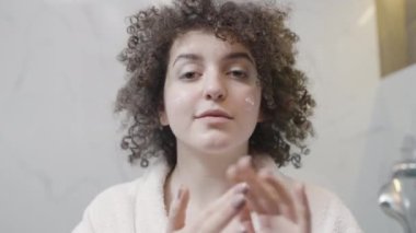 Cilt kremi kullanan gülümseyen genç bir kadın. Aynadaki yansımasından memnun.