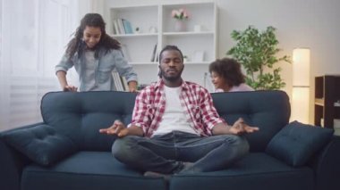 Siyah baba yoga pozisyonunda otururken sakin kalmaya çalışıyor, çocuklar etrafta koşuşturuyor.