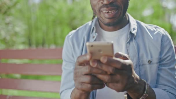 緑の公園のベンチに座っている黒人男性の笑顔 スマートフォンを使用して レジャー — ストック動画