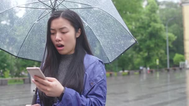 若いアジアの女性は 傘の下で雨の中に立って メッセージを受信 — ストック動画