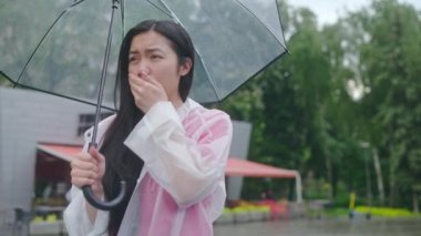 Şemsiyeli genç Asyalı kadın öksürüyor, yağmurda üşüyor, taksi bekliyor.