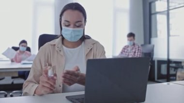 Maskeli kadın programcı dezenfektanla elleri dezenfekte ediyor, dizüstü bilgisayarla çalışıyor.