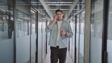 Genç bilişim uzmanı telefonda konuşuyor, ofiste dizüstü bilgisayarla yürüyor, iş yapıyor.