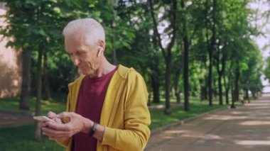 50 'li yaşlarda bir adam akıllı telefondan mesaj yazıyor, parkta müzik dinliyor.