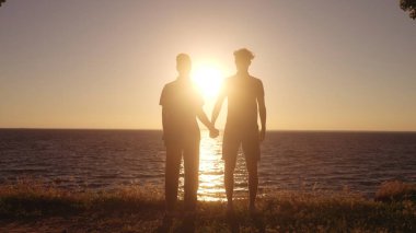 Birbirlerine aşık iki adam el ele tutuşup kumsalda gün batımının tadını çıkarıyorlar.