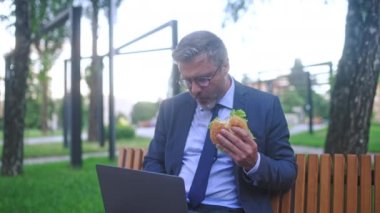 Gözlüklü bir iş adamı laptopla çalışıyor ve dışarıda hamburger yiyor, fast food yiyor.
