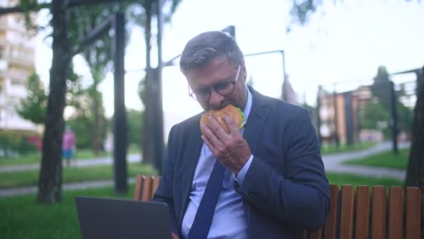 Mężczyzna Garniturze Jedząc Hamburgera Podczas Pracy Laptopie Zewnątrz Odczuwając Ból — Wideo stockowe