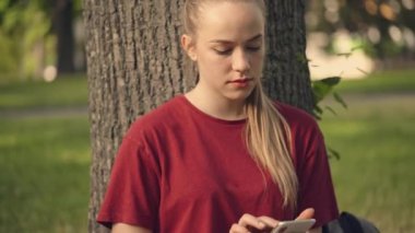 Genç bir kadın akıllı telefondan flört uygulamasını kaydırıyor, parktaki ağacın altında oturuyor.