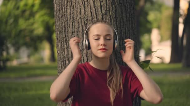 ヘッドフォンで好きな音楽を楽しむ若い女性 公園で木の下に座って — ストック動画