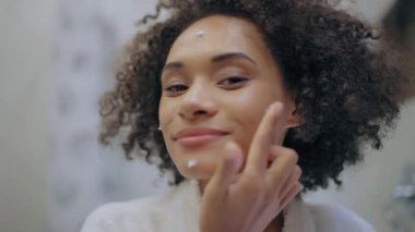 Mutlu genç bayan yüzüne nemlendirici krem sürüyor, kendine bakım rutini, yaş