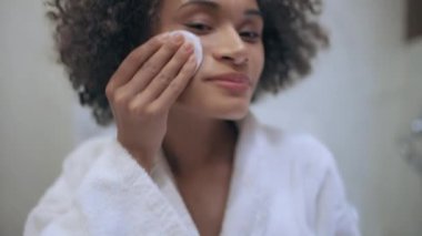 Tatmin olmuş siyah kadın yüzünü losyonla temizliyor. Bakıyor, görünüşünü değiştiriyor.