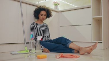 Mutfak zemininde oturan, temizlik deterjanlarına bakan üzgün genç kadın.