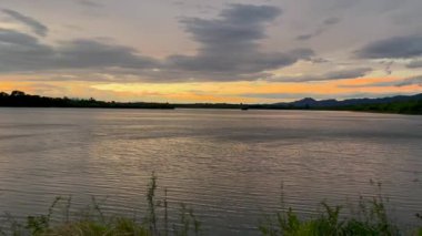 Suyun üzerinde parlak turuncu yansımaları olan sakin bir nehrin üzerinde altın gün batımı. Siluetli ağaçlar ve ön planda yabani otlar alacakaranlıkta huzurlu ve huzurlu bir manzara yaratır.