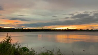 Suyun üzerinde parlak turuncu yansımaları olan sakin bir nehrin üzerinde altın gün batımı. Siluetli ağaçlar ve ön planda yabani otlar alacakaranlıkta huzurlu ve huzurlu bir manzara yaratır.