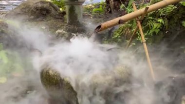 Su bambudan akar, yemyeşil bir bahçede serin bir taş leğen bambudan damlayan su, yükselen yoğun buharla çevrili, doğal bir açık havada gizemli, sakin bir atmosfer yaratır.