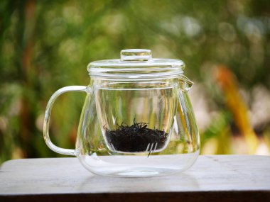 İçinde siyah çay yaprakları olan temiz cam çaydanlık, doğal yeşil bokeh arka planlı ahşap masanın üzerine yerleştirilmiş. Asgari çay demleme konsepti, sakin yaşam tarzı, sağlıklı içecek, temiz tasarım, rahatlama