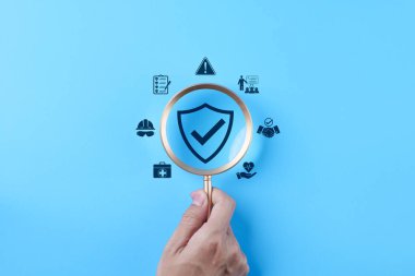 İş güvenliği konsepti. İş sağlığı, risk yönetimi, uyum politikası, endüstriyel koruma, iş yeri güvenlik standartlarını temsil eden simge üzerinde el ele tutuşan büyüteç