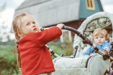 Kız kardeşi, erkek kardeşiyle bebek arabasında oynuyor, bebeği caddede yuvarlıyor.