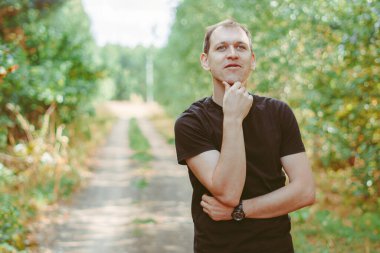 Siyah tişörtlü genç bir adam ağaçların arasında duruyor.