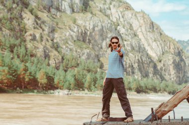 Bir adam dağın arka planında, bir dağ nehrinin yanında poz veriyor.