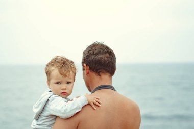 Bir baba oğlunu kollarına alır, denizin yanında durur.