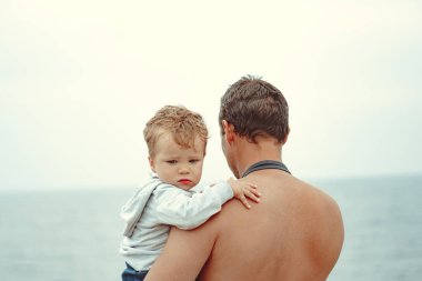 Bir baba oğlunu kollarına alır, denizin yanında durur.