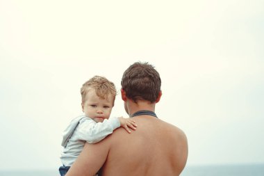 Bir baba oğlunu kollarına alır, denizin yanında durur.