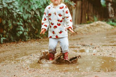 Küçük bir kız lastik botlarla su birikintilerinde koşuyor.