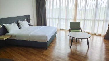 Geniş, modern bir yatak odası, koyu ahşap mobilyalar, beyaz çarşaflı büyük boy bir yatak, ve şık bir orta çağ yeşil koltuğu var..