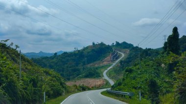 Uzun, dolambaçlı bir asfalt yol yemyeşil tepeler boyunca kıvrılıyor ve bulutlu bir gökyüzü altında tropikal bitki örtüsü.