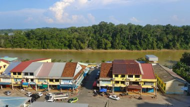 Açık bir gökyüzünün altında yoğun yeşil bir ormanla çevrili tropikal bir nehir kıyısı boyunca geleneksel dükkanların bulunduğu renkli bir kanowit kasabasının yüksek açılı panoramik manzarası.