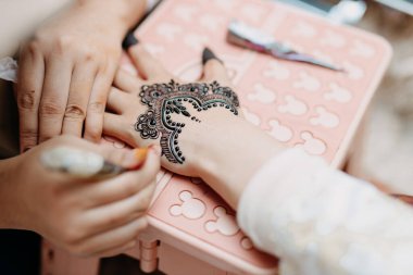 Bir kadının elinin arkasına karmaşık siyah kına (mehendi) desenleri uygulayan bir sanatçı