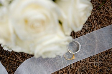 Beyaz kurak çam iğneleri üzerinde alyansların yukarıdan görünüşü, ön planda yumuşak odaklı beyaz bir gül buketi.