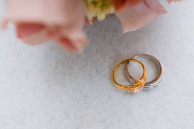 Elmas ve gümüş alyanslı altın bir nişan yüzüğünün yakın plan ayrıntıları yumuşak pembe çiçeklerin yanında beyaz bir yüzey üzerinde dinleniyor.