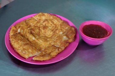 Baharatlı sambal soslu pembe bir tabakta servis edilen bir parça altın-kahverengi Roti Canai (yassı ekmek)..