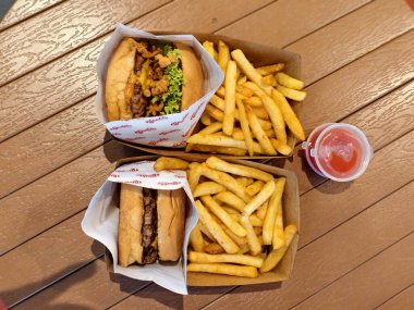Büyük porsiyonlarda altın patates kızartması ve ahşap bir masanın üzerinde kırmızı biber sosuyla servis edilen markalı kâğıt ambalajlı iki gurme sığır burger. 2 Temmuz 2025, Kuching, Sarawak, Malezya