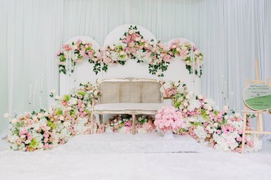 Çiçek aranjmanlı zarif Pastel Düğün Pelamin Dekoru. Beyaz ve pembe güller, bereketli yeşillikler ve örtülü bir arka fona karşı klasik tarzda bir krem kanepe içeren güzel bir Malay düğün kürsüsü (pelamin).