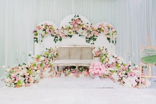 Çiçek aranjmanlı zarif Pastel Düğün Pelamin Dekoru. Beyaz ve pembe güller, bereketli yeşillikler ve örtülü bir arka fona karşı klasik tarzda bir krem kanepe içeren güzel bir Malay düğün kürsüsü (pelamin).
