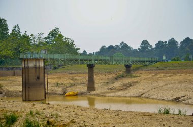 Endüstriyel Su Rezervuarı Köprüsü Kuru Çatlaklı Havza üzerinde. Uzun bir metal servis köprüsü, su seviyesinin önemli ölçüde düşük ve kurumuş, çatlamış toprağı gösteren bir su deposundaki beton giriş kulesine götürür..