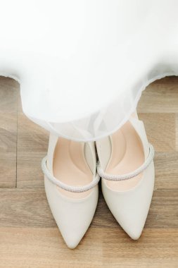 Kristal Straplı zarif Beyaz Burunlu Gelinlik Ayakkabıları. Beyaz bir gelinliğin kenarına takılmış pırıl pırıl bir yapay elmasla süslenmiş, krem renkli, sivri uçlu, düz tabanlı düğün ayakkabıları..