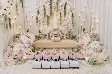 Hantaran Hediyeleriyle Şatafatlı Malezyalı Düğün Pelamin Arkaplanı. Zarif ve süslü bir düğün kürsüsü (Pelamin), ön planda geleneksel paketlenmiş çeyiz kutuları (Hantaran) bulunan, gür beyaz ve pembe çiçeklerle süslenmiş..