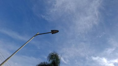 Yumuşak bulutlu açık mavi gökyüzüne karşı alçak bir açıdan çekilmiş modern bir sokak ışığı. Görüntü şehir altyapısı, açık hava aydınlatma ve şehir, fayda ve çevre temaları için uygun minimalist kompozisyonu vurguluyor