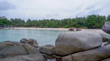 Siangau Sahili 'nin ön planında, sakin bir deniz ve arka planda yoğun hindistan cevizi palmiyesi ormanı bulunan büyük gri granit bir kaya parçası bulunur.