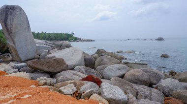 Kızıl toprak zemin, gri granit kayalar ve Endonezya 'da sakin bir deniz ufkunun yer aldığı kayalık bir kıyı şeridi manzarası..