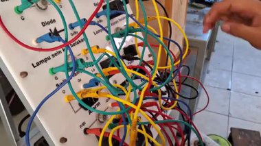 Otomotiv atölyesindeki eğitim panelinde motorsiklet elektrik tesisatı pratiği yapan mesleki öğrencilerin yakın çekimi. Renkli kabloları diyota bağlayan, fitil, sinyal lambasına çeviren eller.